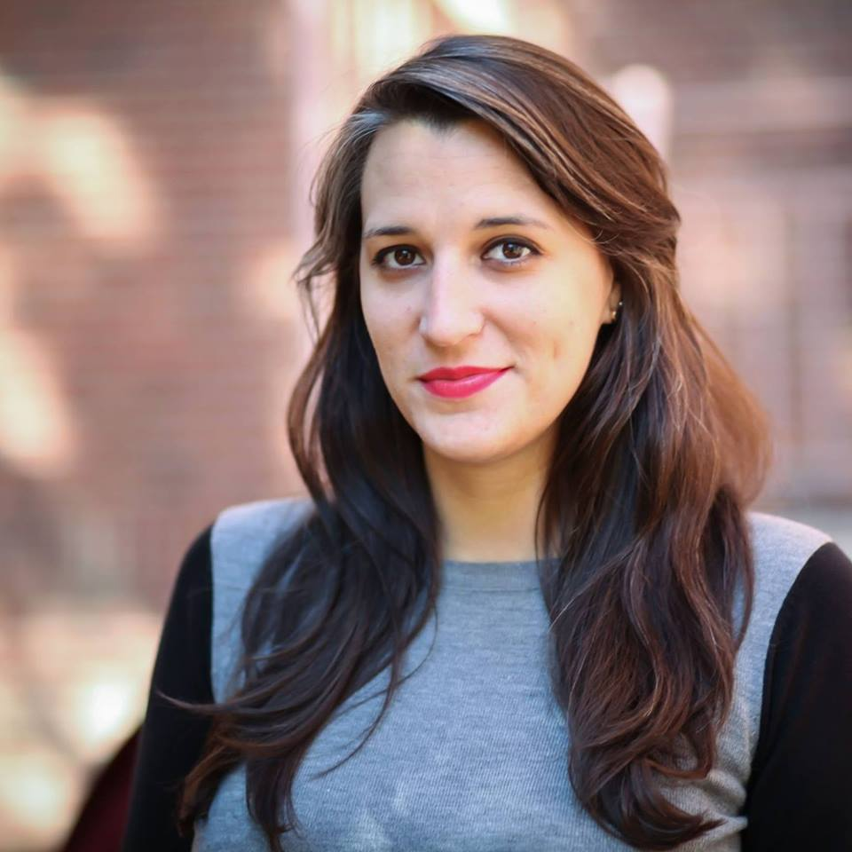 A woman with long brown and gray hair and brown eyes, wearing a black and gray sweater, stands outside and smiles at the camera.